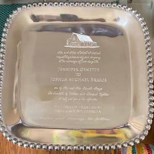 May include: A silver-colored, square serving tray with a beaded edge. The tray is engraved with a barn illustration and wedding details, including names and a location. The text reads "Jennifer Genette to Joshua Michael Briggs".