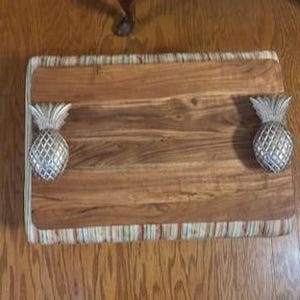 May include: A rectangular wooden serving tray featuring a natural brown wood grain surface and a striped fabric border. The tray is equipped with two silver-colored metal handles shaped like pineapples on the left and right sides.