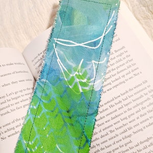 May include: A green and blue fabric bookmark with white painted lines. The bookmark is frayed on the edges and has a stitched seam along the side.