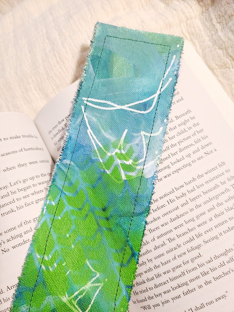 May include: A green and blue fabric bookmark with white painted lines. The bookmark is frayed on the edges and has a stitched seam along the side.