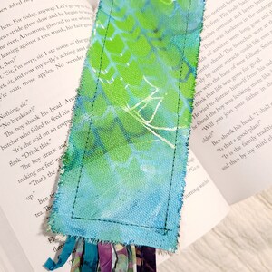 May include: A handmade bookmark with a blue and green watercolor design. The bookmark is made of fabric and has a fringe of fabric at the bottom. The bookmark is placed on a book with text.