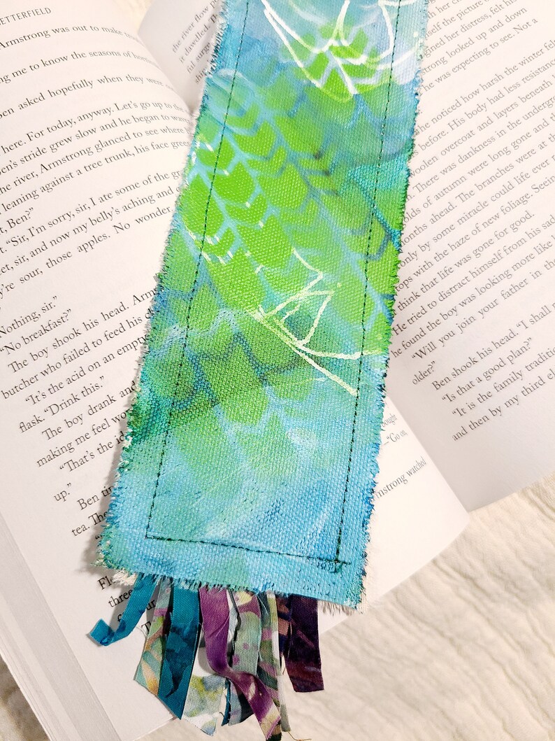May include: A handmade bookmark with a blue and green watercolor design. The bookmark is made of fabric and has a fringe of fabric at the bottom. The bookmark is placed on a book with text.