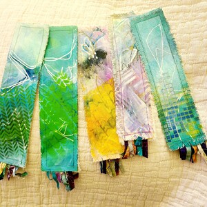 May include: Five handmade fabric bookmarks with frayed edges and colorful abstract designs. Each bookmark has a different color scheme and pattern, including green, blue, yellow, purple, and teal. The bookmarks are decorated with white lines and have colorful fabric tassels.