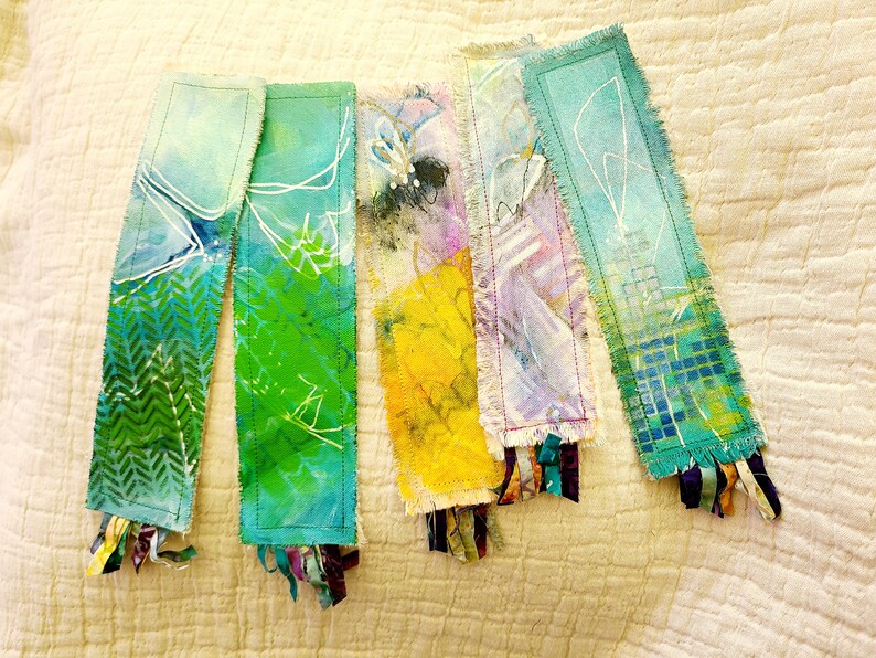 May include: Five handmade fabric bookmarks with frayed edges and colorful abstract designs. Each bookmark has a different color scheme and pattern, including green, blue, yellow, purple, and teal. The bookmarks are decorated with white lines and have colorful fabric tassels.