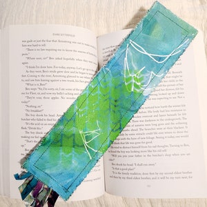 May include: A blue and green fabric bookmark with white painted designs. The bookmark has a frayed edge and a tassel made of fabric scraps.