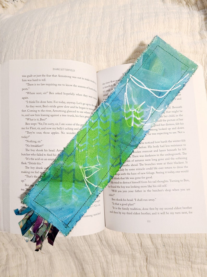 May include: A blue and green fabric bookmark with white painted designs. The bookmark has a frayed edge and a tassel made of fabric scraps.