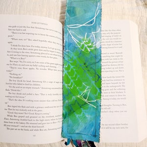 May include: A close-up of a book with a blue and green fabric bookmark. The bookmark has a white design on it. The book is open to a page with text about a boy who ate a green apple and got sick.