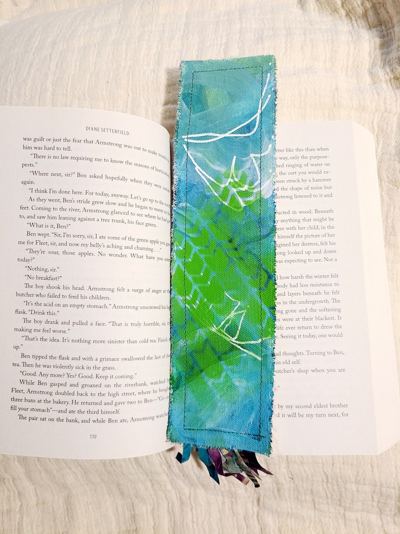 May include: A close-up of a book with a blue and green fabric bookmark. The bookmark has a white design on it. The book is open to a page with text about a boy who ate a green apple and got sick.