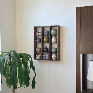 May include: A wooden display shelf with a grid of sixteen compartments, each holding a small ceramic vessel. The vessels vary in shape, color, and design, including vases, mugs, and decorative pieces. The shelf is mounted on a white wall.