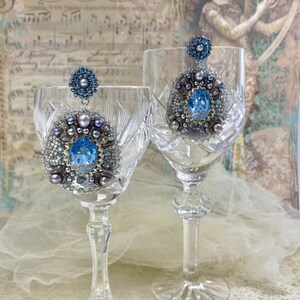 May include: A pair of silver and blue beaded earrings with a large blue gemstone in the center of each earring. The earrings are hanging from two crystal wine glasses.