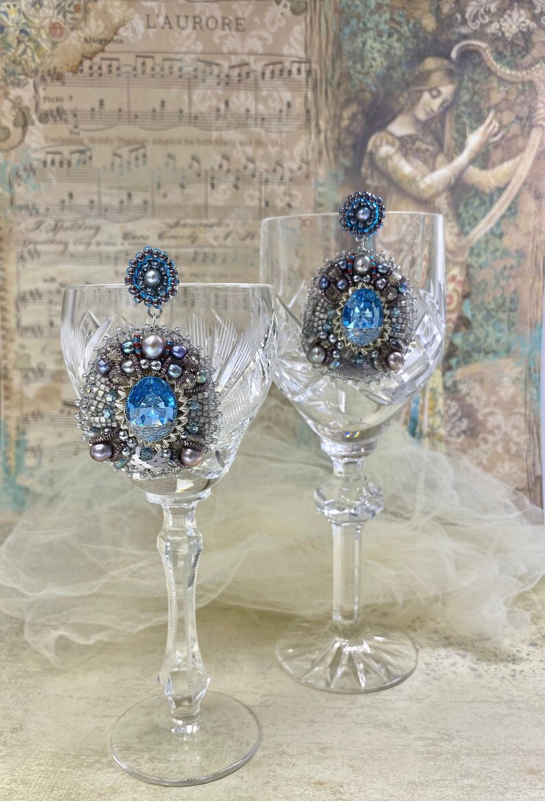May include: A pair of silver and blue beaded earrings with a large blue gemstone in the center of each earring. The earrings are hanging from two crystal wine glasses.