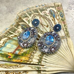 May include: A pair of silver and blue beaded earrings with a large blue gemstone in the center of each earring. The earrings are displayed on a vintage fan.