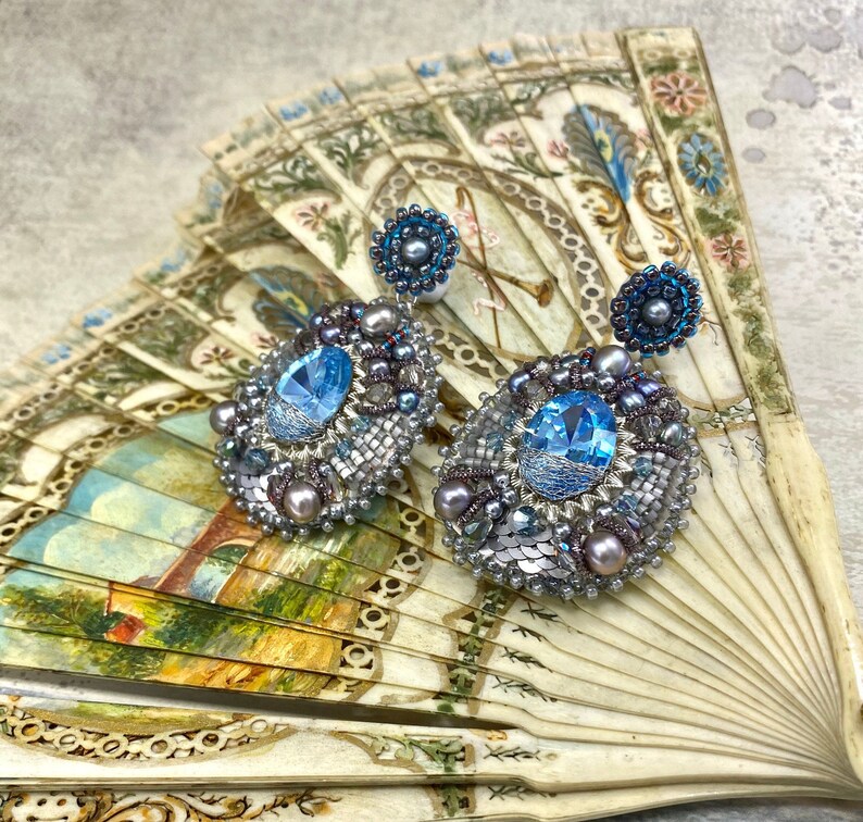 May include: A pair of silver and blue beaded earrings with a large blue gemstone in the center of each earring. The earrings are displayed on a vintage fan.