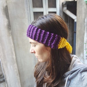 May include: A purple and yellow knitted headband with a long yellow tassel. The headband is worn by a person with long brown hair.