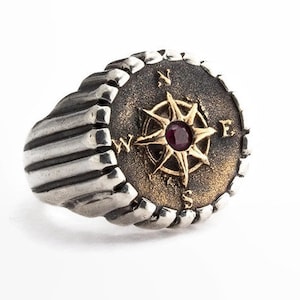 May include: A silver ring with a compass design. The ring features a circular face with a gold compass rose and a red gemstone center. The band has a ribbed design. The compass points to the cardinal directions.