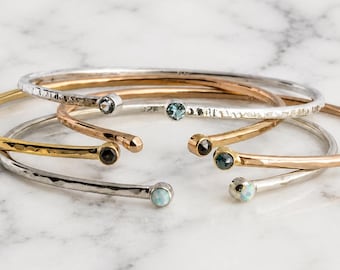 May include: A collection of four open-ended cuff bracelets featuring hammered metal bands in silver, gold, and rose gold tones. Each bracelet is finished with a small, round, bezel-set gemstone at both ends, including blue, white, and dark stones.