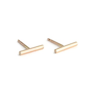 May include: A pair of gold-colored bar stud earrings. Each earring has a straight bar design with a post for piercing. The earrings are set against a white background.