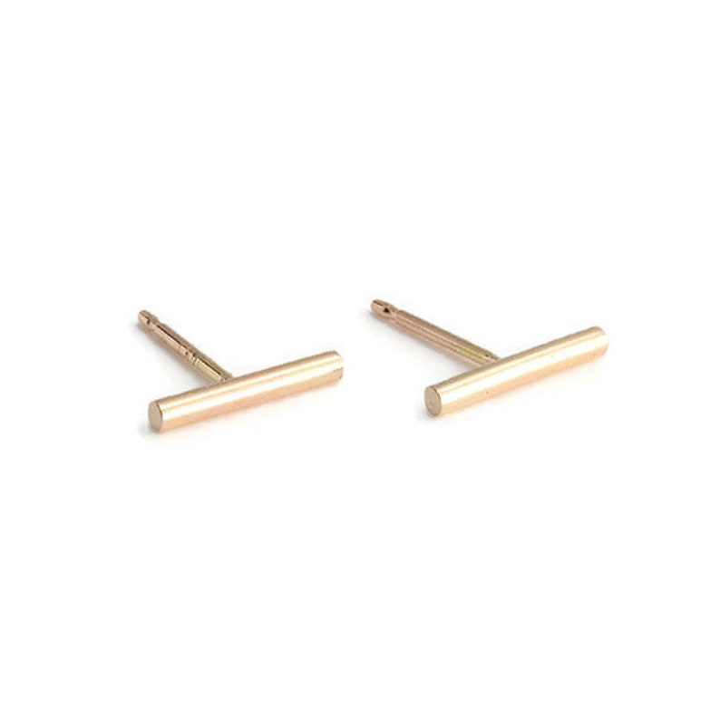 May include: A pair of gold-colored bar stud earrings. Each earring has a straight bar design with a post for piercing. The earrings are set against a white background.