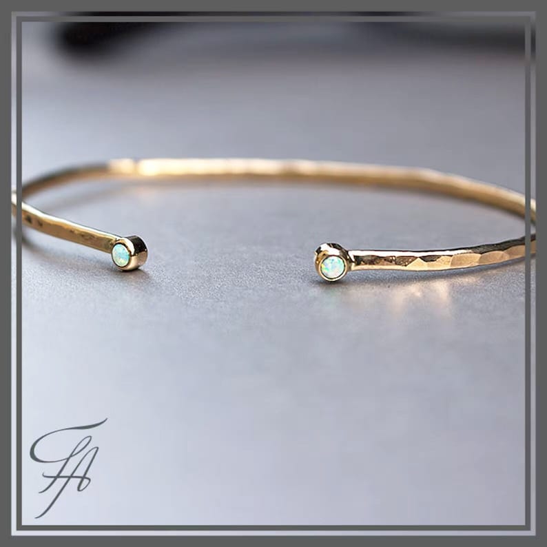 Pode incluir: Uma pulseira de punho aberta dourada com textura martelada. Cada extremidade da pulseira apresenta uma pequena pedra preciosa redonda e iridescente. A pulseira &eacute; exibida em um fundo neutro, destacando sua eleg&acirc;ncia simples.