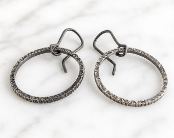 Pode incluir: Um par de brincos de argola em metal cinza-escuro com um acabamento texturizado e martelado. Cada argola circular está suspensa por um gancho de orelha em metal cinza-escuro a condizer, criando um design simples e coerente.