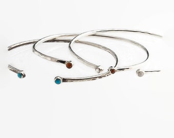Pode incluir: Uma coleção de pulseiras de punho de metal prateado. Cada pulseira apresenta um design liso e curvo com um pequeno detalhe de pedra colorida na extremidade. As pedras são de várias cores, incluindo turquesa, castanho e branco.