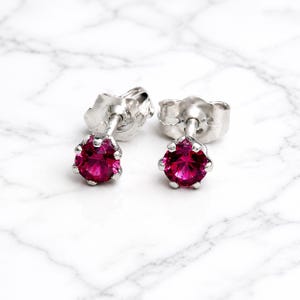 May include: A pair of silver stud earrings featuring bright, round, magenta gemstones. The earrings have a classic design, with the gemstones set in a prong setting. The posts and backs are also silver. The earrings are displayed on a white marble surface.