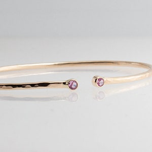Pode incluir: Uma pulseira de punho aberta dourada com textura martelada. A pulseira apresenta uma pequena pedra preciosa rosa redonda em cada extremidade aberta. A joia &eacute; exibida em uma superf&iacute;cie branca.