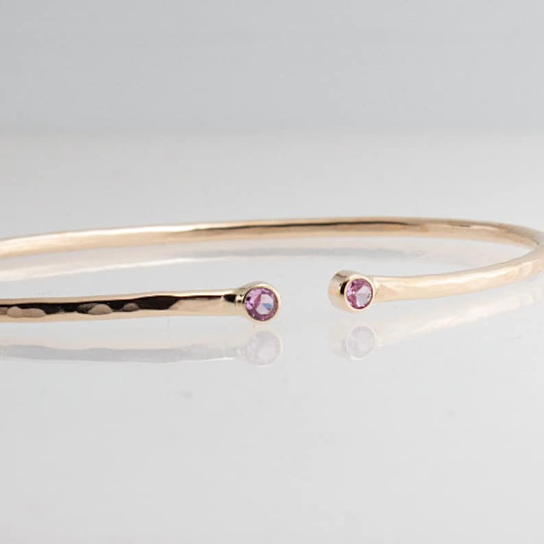 Pode incluir: Uma pulseira de punho aberta dourada com textura martelada. A pulseira apresenta uma pequena pedra preciosa rosa redonda em cada extremidade aberta. A joia &eacute; exibida em uma superf&iacute;cie branca.