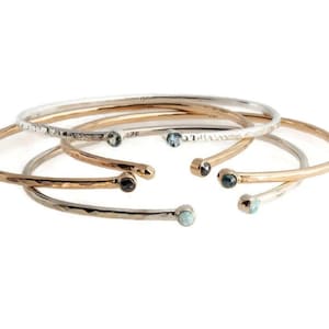 Pode incluir: Uma cole&ccedil;&atilde;o de pulseiras abertas em tons de prata, ouro e ouro rosa. Cada pulseira apresenta uma textura martelada e &eacute; adornada com uma pequena pedra preciosa em cada extremidade. As pedras preciosas incluem pedras azuis, pretas e brancas.