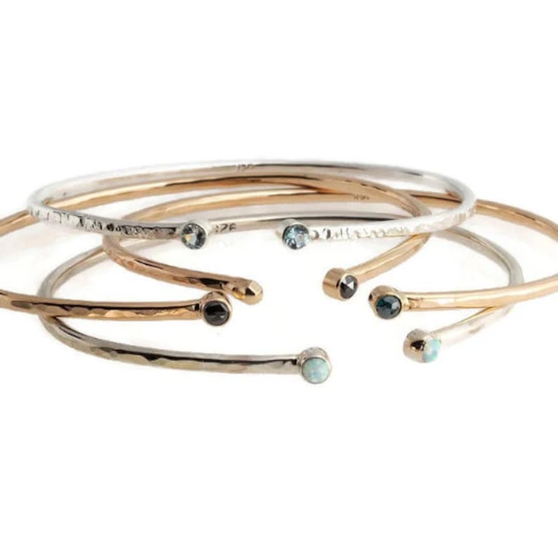 Pode incluir: Uma cole&ccedil;&atilde;o de pulseiras abertas em tons de prata, ouro e ouro rosa. Cada pulseira apresenta uma textura martelada e &eacute; adornada com uma pequena pedra preciosa em cada extremidade. As pedras preciosas incluem pedras azuis, pretas e brancas.