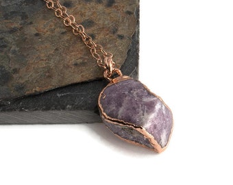 May include: A rose gold necklace featuring a raw, irregular-shaped, purple gemstone pendant. The pendant is encased in a rose gold setting and hangs from a delicate chain. The gemstone has a natural, textured appearance.