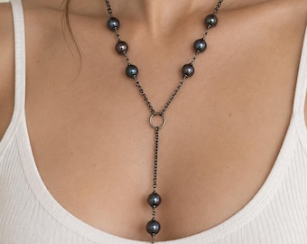 May include: A lariat-style necklace featuring dark, iridescent pearls strung along a blackened metal chain. The design includes a central circular ring connector with a vertical drop section ending in a small, coiled metal snake charm.