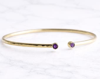 May include: A gold-colored open cuff bracelet with a hammered texture. The bracelet features a small, round, purple gemstone at each open end. The jewelry is displayed on a white marble surface.