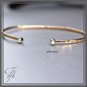 Pode incluir: Uma pulseira de punho aberta dourada com textura martelada. Cada extremidade da pulseira apresenta uma pequena pedra de opala branca redonda. As palavras "white Opal" s&atilde;o impressas em uma fonte cursiva.