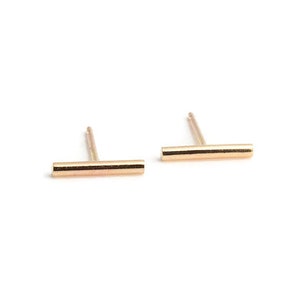 May include: A pair of gold-colored bar stud earrings. Each earring features a straight, horizontal bar connected to a vertical post. The earrings are set against a white background.