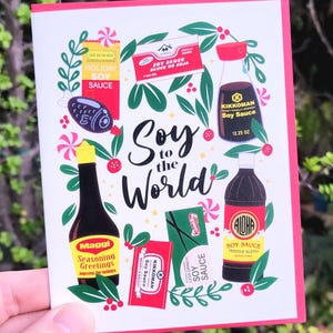 May include: A greeting card with a pink border featuring various soy sauce bottles and packages, with the text "Soy to the World." The card includes illustrations of soy sauce brands like Kikkoman and Aloha, and the text "Holiday Soy Sauce."