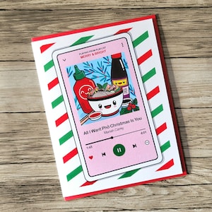 May include: A greeting card with a pink phone screen design featuring a bowl of pho, hot sauce, and soy sauce. The text reads "All I Want Phở Christmas Is You" and "Merry & Bright" on a red and white striped background.