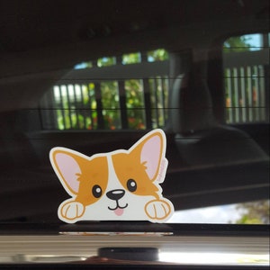 May include: A sticker of a cartoon corgi dog peeking over the edge of a car window. The corgi is orange and white with pink ears and a pink tongue sticking out. The sticker has the text "Corgi Vibes" on the side.
