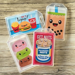 May include: Four clear plastic badge holders with colorful cartoon images inside. The images are of a hamburger, boba tea, mochi, and macadamia nuts. The text on the badge holders reads "Bun Voyage!", "Get Nuts", "Macadamia Nuts Made in Hawaii", and "Get Nuts".