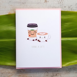 May include: A white greeting card with a pink border features a cartoon illustration of a coffee cup and a to-go coffee cup with smiling faces and coffee beans. The text "THANKS A LATTE" is printed below the illustrations.