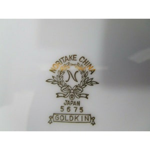 Op de afbeelding: Een gouden Noritake China logo met een krans en de letter N in het midden. Het logo bevat ook de tekst "Japan 5675 Goldkin".