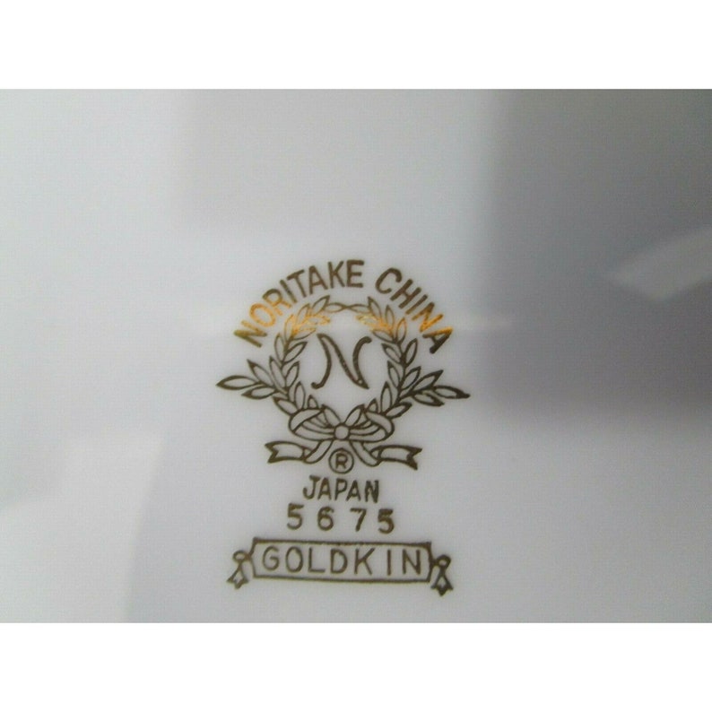 Op de afbeelding: Een gouden Noritake China logo met een krans en de letter N in het midden. Het logo bevat ook de tekst "Japan 5675 Goldkin".