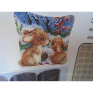 May include: A small, square needlepoint pillow featuring two brown bunnies in a snowy scene with blue and green foliage and red berries. The bunnies are facing each other, with one appearing to nuzzle the other. The pillow has a white border.
