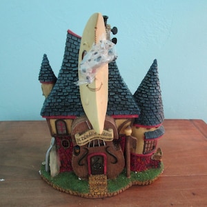 May include: A whimsical decorative house figurine with a violin-shaped facade. The house features a smiling moon, a cello, and the text "H. Diddle Diddles". The roof is dark blue, and the walls are red and yellow.