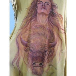 Puede incluir: Un chaleco de cuero marr&oacute;n con una imagen pintada de una mujer con el pelo largo y suelto y un b&uacute;falo. El b&uacute;falo est&aacute; mirando hacia adelante con la cabeza baja. Los ojos de la mujer est&aacute;n cerrados y ella est&aacute; mirando hacia arriba. El chaleco tiene flecos alrededor de los bordes.