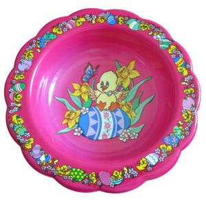 Può includere: Una ciotola rosa brillante con bordo smerlato a tema pasquale. La ciotola presenta una grafica centrale di un pulcino giallo su un uovo blu decorato, circondato da fiori e una farfalla. Il bordo è decorato con uova e pulcini pasquali.