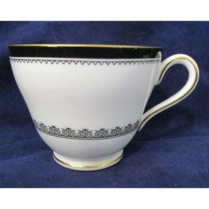 Puede incluir: Una taza de té blanca con un borde negro y dorado. La taza tiene un diseño negro y dorado alrededor del borde y un diseño negro y dorado alrededor de la base.