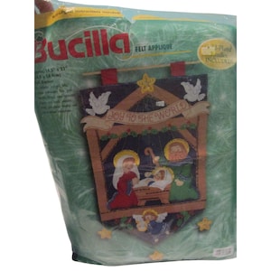 May include: A Bucilla felt applique kit featuring a Christmas nativity scene. The banner includes the text "Joy to the World" and images of Mary, Joseph, baby Jesus, angels, and stars. The package is 14.5" x 23".