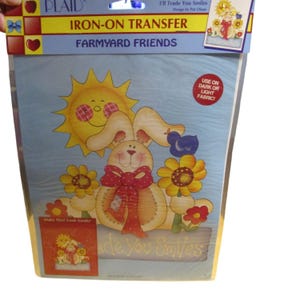 May include: A Plaid brand iron-on transfer package titled Farmyard Friends featuring a cartoon rabbit with a red polka dot bow, a yellow sun, sunflowers, and a blue bird. The packaging includes the text I'll Trade You Smiles and Use on Dark or Light Fabric.