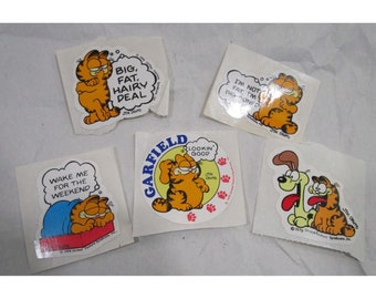 May include: Five Garfield comic strip stickers featuring the orange cat Garfield. The stickers feature different sayings, including "Wake me for the weekend", "Big, Fat, Hairy Deal", "Lookin' Good", "I'm not fat I'm big-boned", and "Garfield" with a dog.